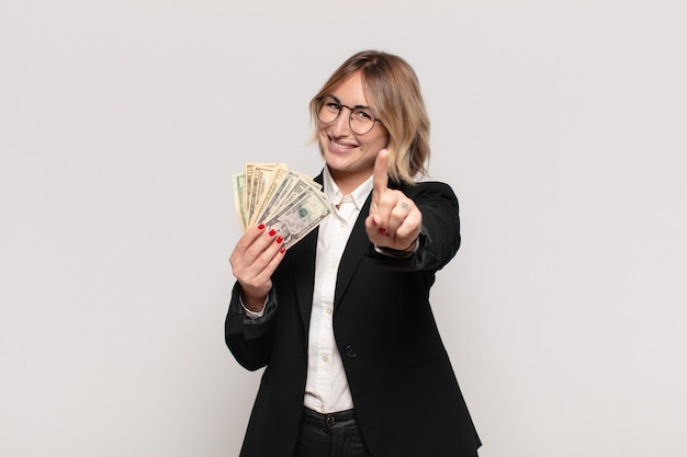 Joven mujer rubia sonriendo con orgullo y confianza haciendo la pose número uno triunfalmente, sintiéndose como una líder