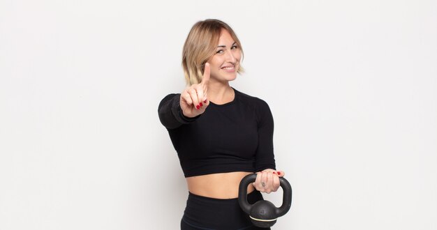 Joven mujer rubia sonriendo con orgullo y confianza haciendo la pose número uno triunfalmente, sintiéndose como una líder