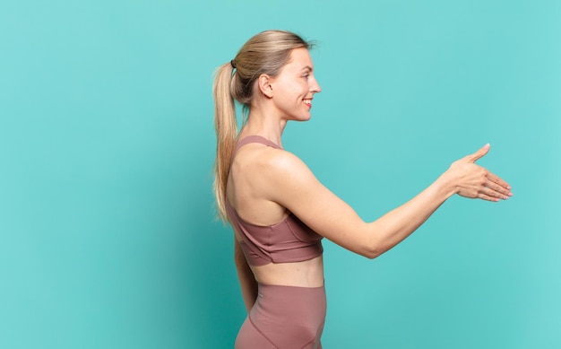 joven mujer rubia sonriendo, saludándote y ofreciendo un apretón de manos para cerrar un trato exitoso, concepto de cooperación. concepto de deporte