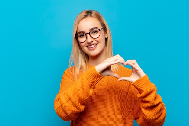 Joven mujer rubia sonriendo y sintiéndose feliz, linda, romántica y enamorada, haciendo forma de corazón con ambas manos