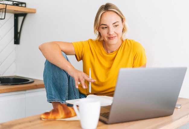 Joven mujer rubia sonriente con camiseta amarilla trabajando en una laptop en la cocina de casa