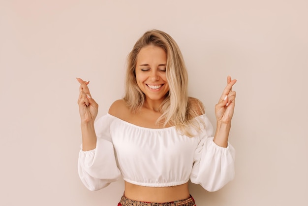 Joven mujer rubia sonriente cruza los dedos para tener buena suerte esperando los resultados de la lotería