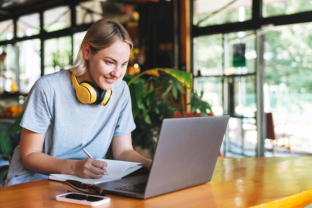 Joven mujer rubia sonriente independiente con auriculares amarillos trabajando en una computadora portátil en el café