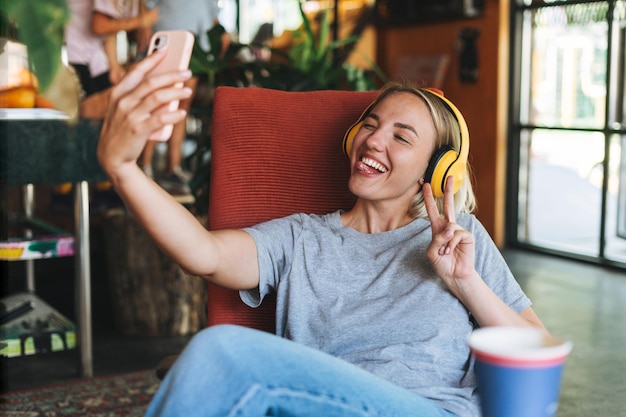 Joven mujer rubia sonriente con los ojos cerrados en auriculares amarillos disfruta de la música con el teléfono móvil en la silla del café Chica feliz tomando selfie en el teléfono inteligente