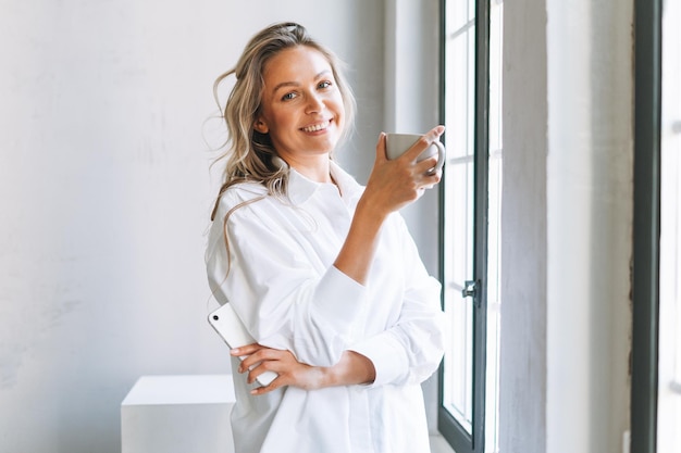 Joven mujer rubia sonriente con el pelo largo en camisa blanca descansa y bebe té cerca de la ventana en la luminosa oficina moderna