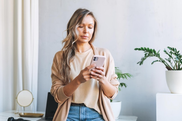 Joven mujer rubia sonriente con el pelo largo en un elegante cárdigan que trabaja en una laptop en la luminosa oficina moderna mujer envía un mensaje por teléfono inteligente