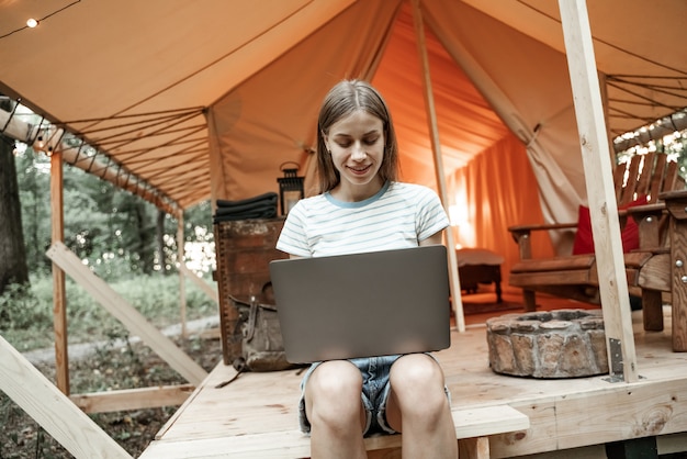 Joven mujer rubia sonriente sentada en suelo glamping escribiendo en la computadora portátil, streaming, mensajería. Estilo de vida de camping. Viajes de bajo presupuesto. Concepto de trabajo remoto. Tecnología moderna de comunicación por internet.