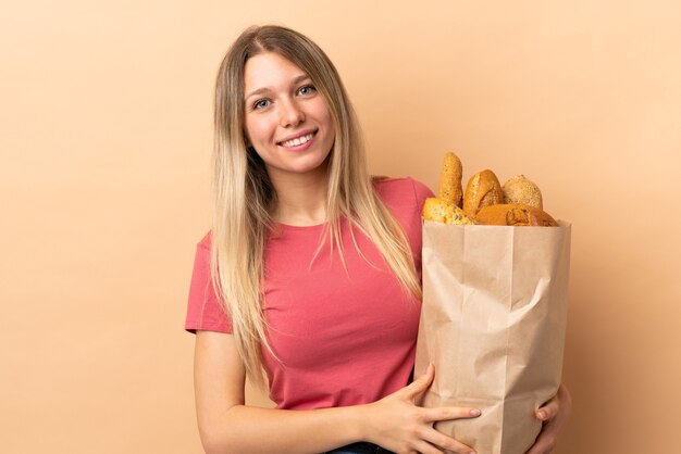 Joven mujer rubia sosteniendo una bolsa llena de panes aislado en beige riendo