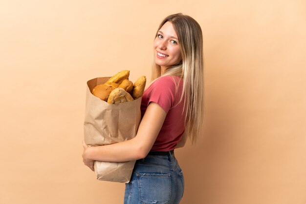 Joven mujer rubia sosteniendo una bolsa llena de panes aislados en la pared beige riendo