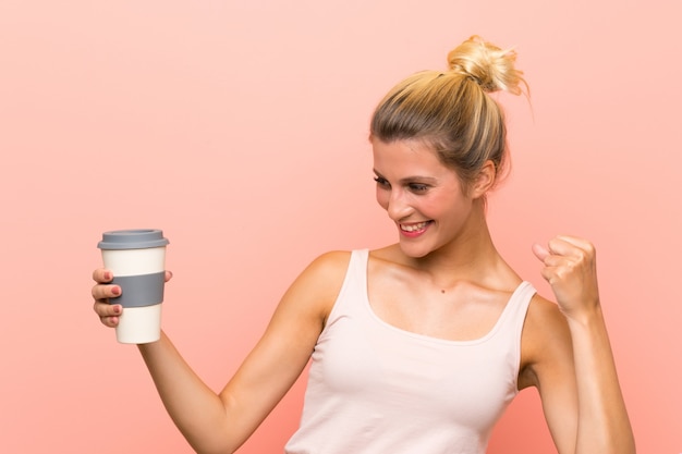 Joven mujer rubia sosteniendo un café para llevar celebrando una victoria