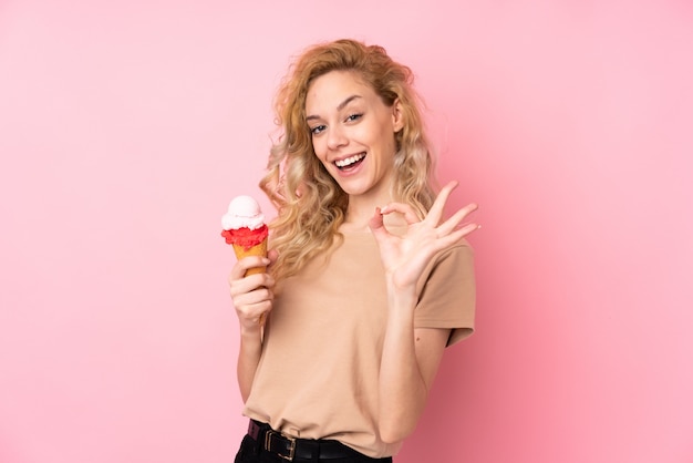 Joven mujer rubia sosteniendo un helado de cucurucho aislado en la pared de color rosa que muestra bien firmar con los dedos