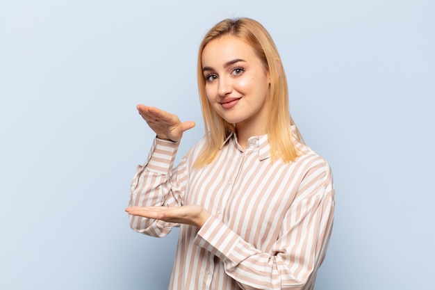 Joven mujer rubia sosteniendo un objeto con ambas manos en el espacio de copia lateral, mostrando, ofreciendo o publicitando un objeto