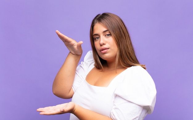 Joven mujer rubia sosteniendo un objeto con ambas manos en el espacio de copia lateral, mostrando, ofreciendo o publicitando un objeto