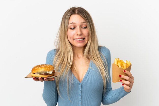 Joven mujer rubia sosteniendo patatas fritas y hamburguesa con queso sobre antecedentes aislados