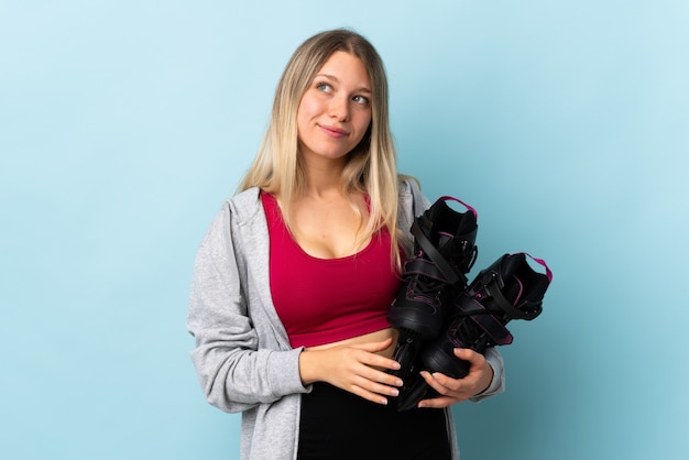 Joven mujer rubia sosteniendo un patines aislado en la pared de color rosa de pie y mirando hacia el lado