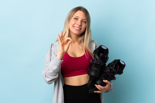 Joven mujer rubia sosteniendo un patines aislados en la pared rosa mostrando un signo de ok con los dedos