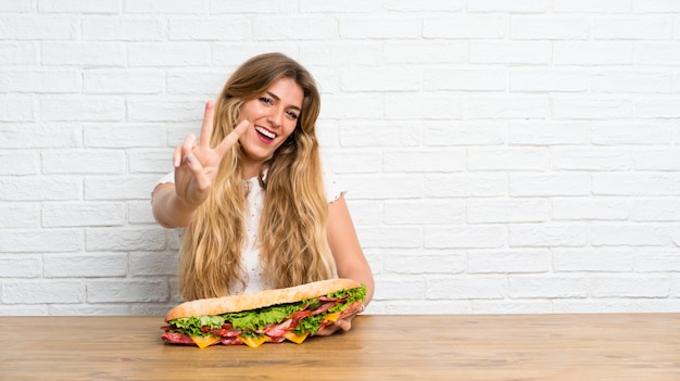 Foto joven mujer rubia sosteniendo un sándwich grande haciendo gesto de victoria