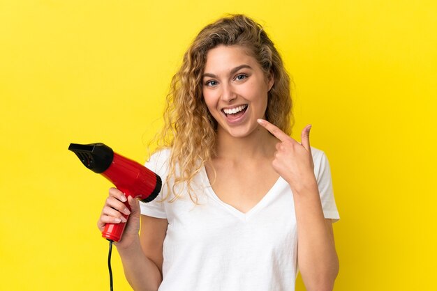 Joven mujer rubia sosteniendo un secador de pelo aislado sobre fondo amarillo dando un pulgar hacia arriba gesto