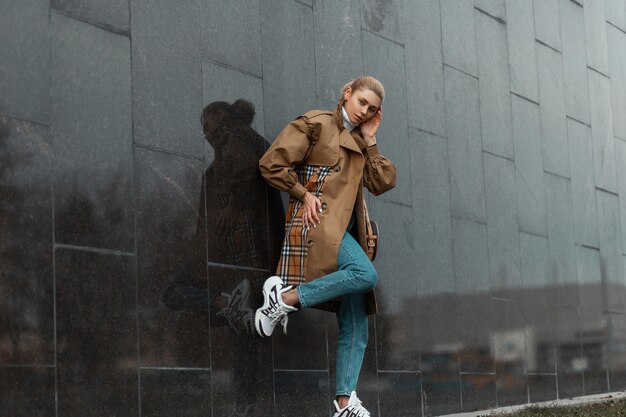 Joven mujer rubia con un suéter y una gabardina beige posando cerca de un edificio moderno negro