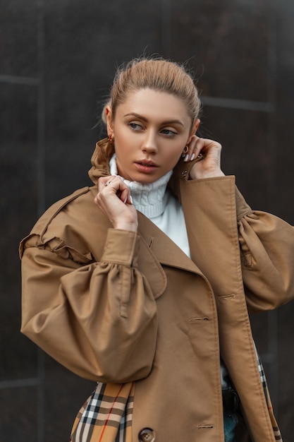 Joven mujer rubia con un suéter y una gabardina beige posando cerca de un edificio moderno negro