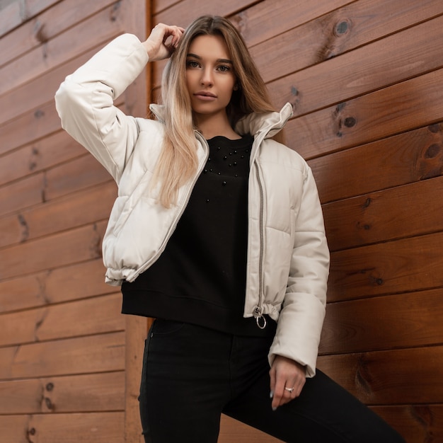 Joven mujer rubia con un suéter negro y una chaqueta blanca de moda posando junto a un edificio de madera