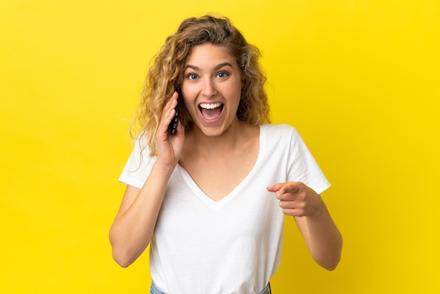 Joven mujer rubia con teléfono móvil aislado sobre fondo amarillo sorprendido y apuntando hacia el frente