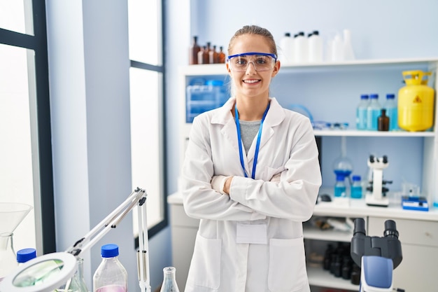 Joven mujer rubia con uniforme científico de pie con gesto de brazos cruzados en el laboratorio