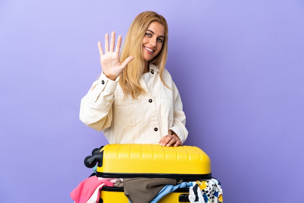 Joven mujer rubia uruguaya con una maleta llena de ropa sobre la pared púrpura aislada contando cinco con los dedos