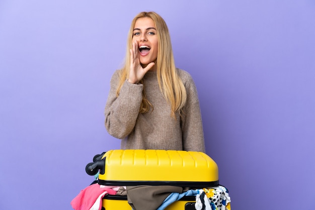 Joven mujer rubia uruguaya con una maleta llena de ropa sobre la pared púrpura aislada gritando con la boca abierta