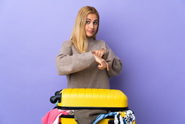 Joven mujer rubia uruguaya con una maleta llena de ropa sobre pared púrpura aislada haciendo el gesto de llegar tarde