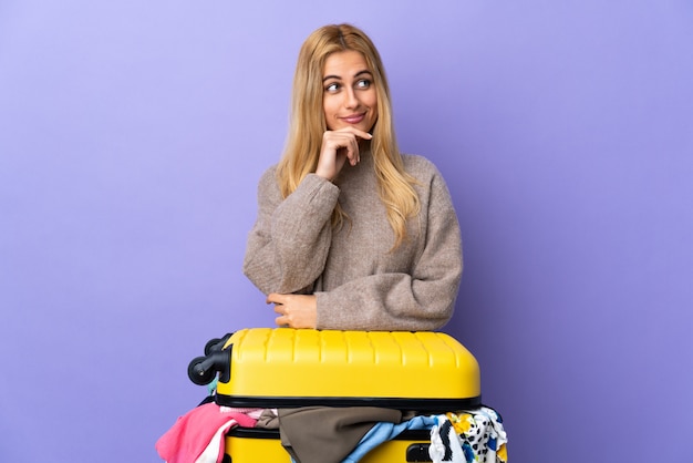 Joven mujer rubia uruguaya con una maleta llena de ropa sobre pared púrpura aislada teniendo dudas y pensando