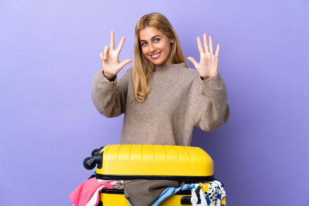 Joven mujer rubia uruguaya con una maleta llena de ropa sobre la pared púrpura contando ocho con los dedos