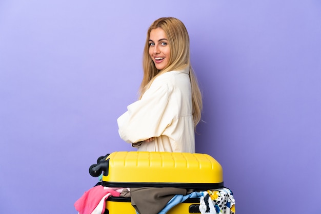 Joven mujer rubia uruguaya con una maleta llena de ropa sobre pared púrpura en posición lateral