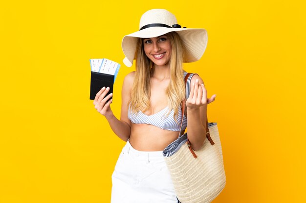 Joven mujer rubia uruguaya en traje de baño sosteniendo un pasaporte sobre fondo amarillo aislado que invita a venir con la mano. Feliz de que hayas venido