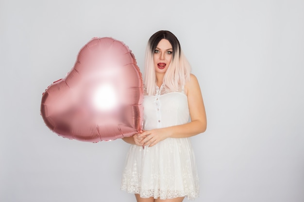 Joven mujer rubia en vestido de encaje blanco sosteniendo un gran globo de corazón rosa en sus manos sobre fondo blanco.