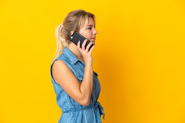 Joven mujer rusa aislada en la pared amarilla manteniendo una conversación con el teléfono móvil con alguien