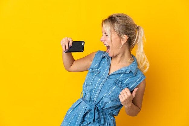 Joven mujer rusa aislada sobre fondo amarillo con teléfono móvil y cantando