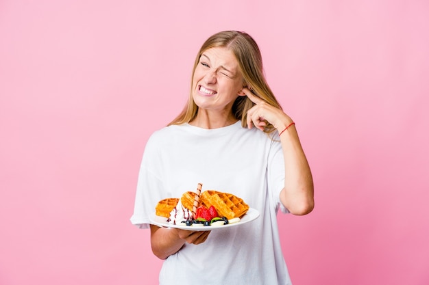 Joven mujer rusa comiendo un gofre aislado cubriendo las orejas con las manos.