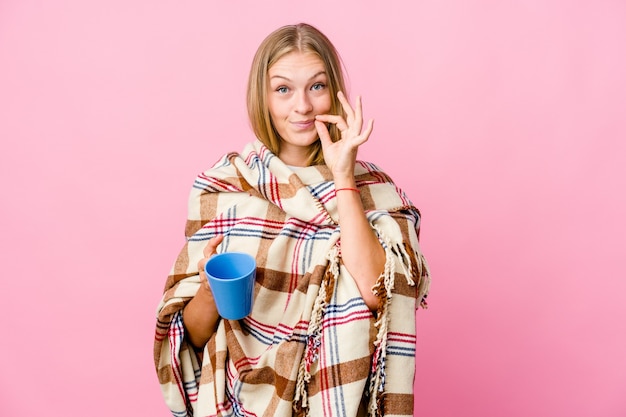 Joven mujer rusa envuelta en una manta bebiendo café con los dedos en los labios guardando un secreto