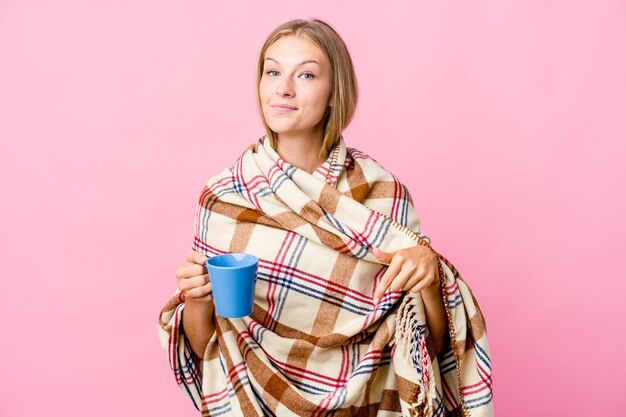 Joven mujer rusa envuelta en una manta tomando café apunta hacia abajo con los dedos, sentimiento positivo