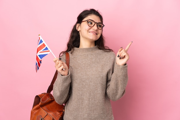 Joven mujer rusa sosteniendo una bandera del Reino Unido aislada en la pared rosa apuntando hacia una gran idea