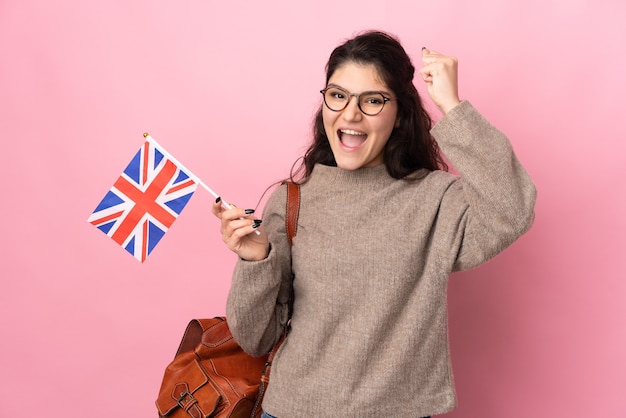 Joven mujer rusa sosteniendo una bandera del Reino Unido aislada en la pared rosa celebrando una victoria