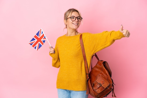 Joven mujer rusa sosteniendo una bandera de Reino Unido aislada en la pared rosa dando un pulgar hacia arriba gesto