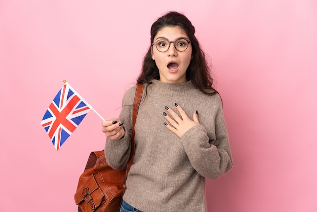 Joven mujer rusa sosteniendo una bandera del Reino Unido aislada sobre fondo rosa con expresión facial sorpresa