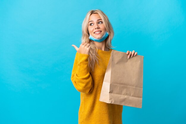 Joven mujer rusa sosteniendo una bolsa de compras aislada en azul apuntando hacia el lado para presentar un producto