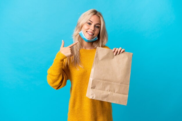 Joven mujer rusa sosteniendo una bolsa de compras aislada en azul dando un pulgar hacia arriba gesto