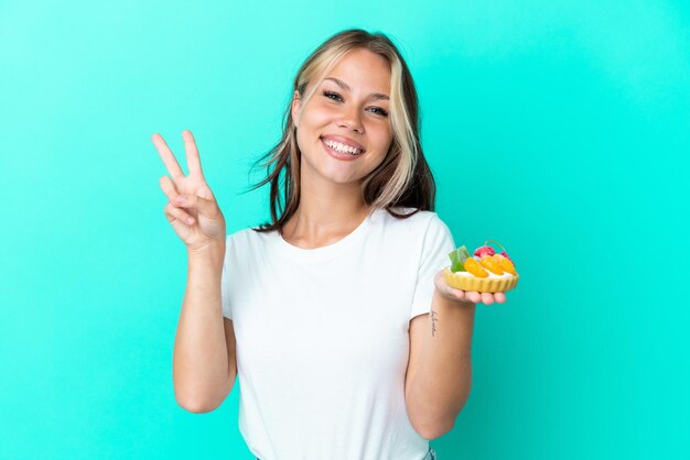 Joven mujer rusa sosteniendo un dulce de frutas aislado sobre fondo azul sonriendo y mostrando el signo de la victoria