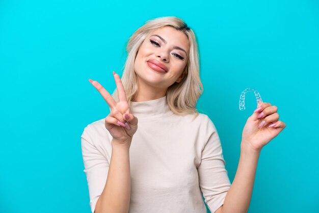 Joven mujer rusa sosteniendo invising aislado sobre fondo azul sonriendo y mostrando el signo de la victoria