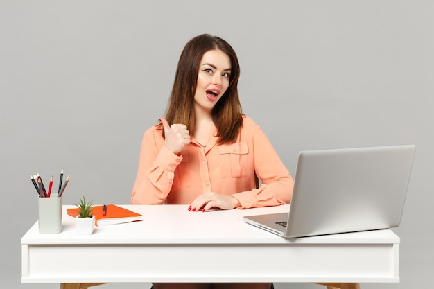 Joven mujer segura de sí misma con ropa informal en colores pastel que muestra el pulgar arriba sentada, trabaja en un escritorio blanco con una computadora portátil aislada en un fondo gris. Concepto de estilo de vida de carrera empresarial de logro. Simulacros de espacio de copia.