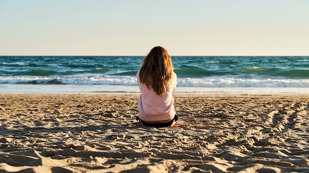 Joven mujer sentada en la arena de la playa, viendo el atardecer